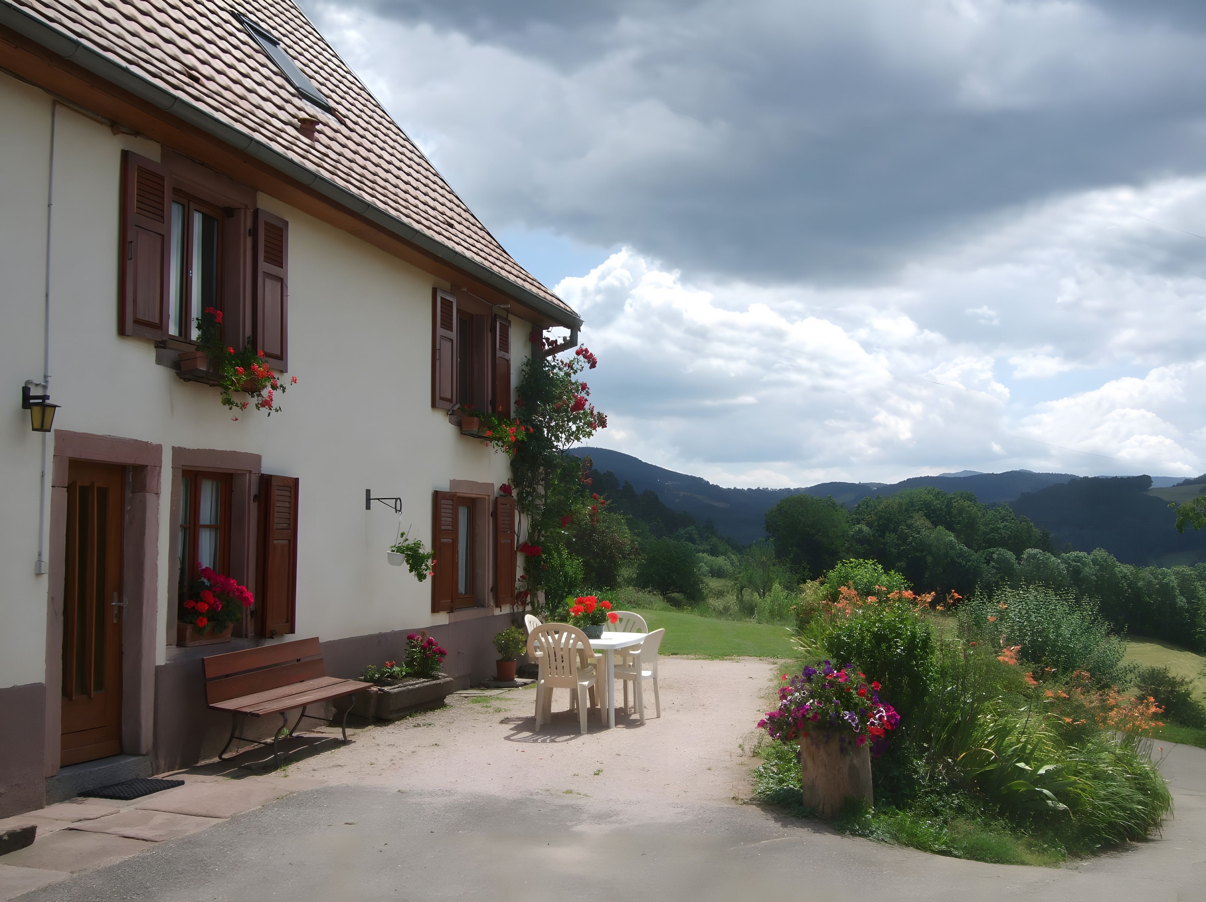 A building with a sloped roof, wooden shutters, and flower boxes. There is a bench and a table with chairs outside.