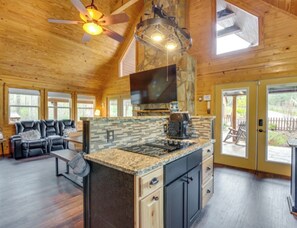 A spacious cabin interior with a large kitchen island, wooden floors, a ceiling fan, and a stone fireplace.