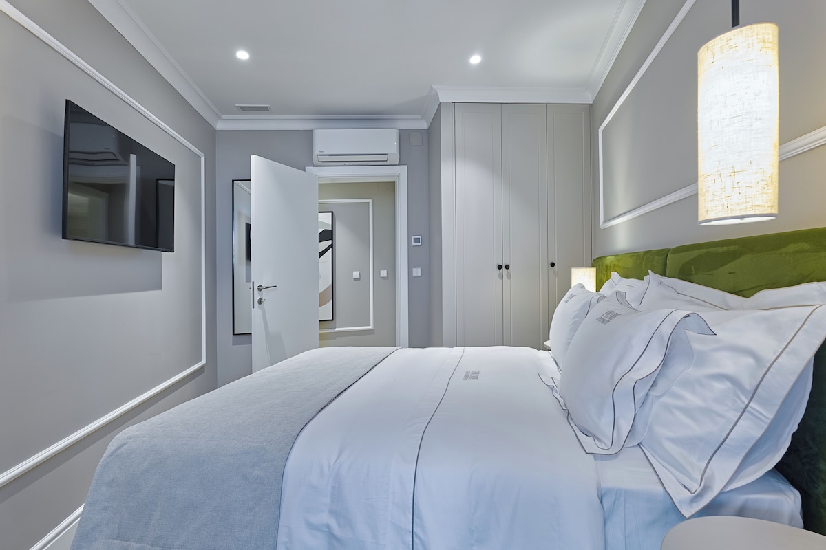 Una habitación de hotel moderna con una cama grande, un televisor de pantalla plana montado en la pared, una lámpara de techo y una puerta que conduce a otra habitación.