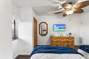 Une chambre à coucher comprenant un lit, une commode, un ventilateur de plafond, un téléviseur et une fenêtre avec des stores.