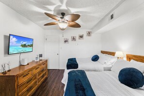 A hotel room with a ceiling fan, a television, a wooden dresser, two beds with blue pillows, and artwork on the walls. - Jamaica Daybreak North (Ocean City)