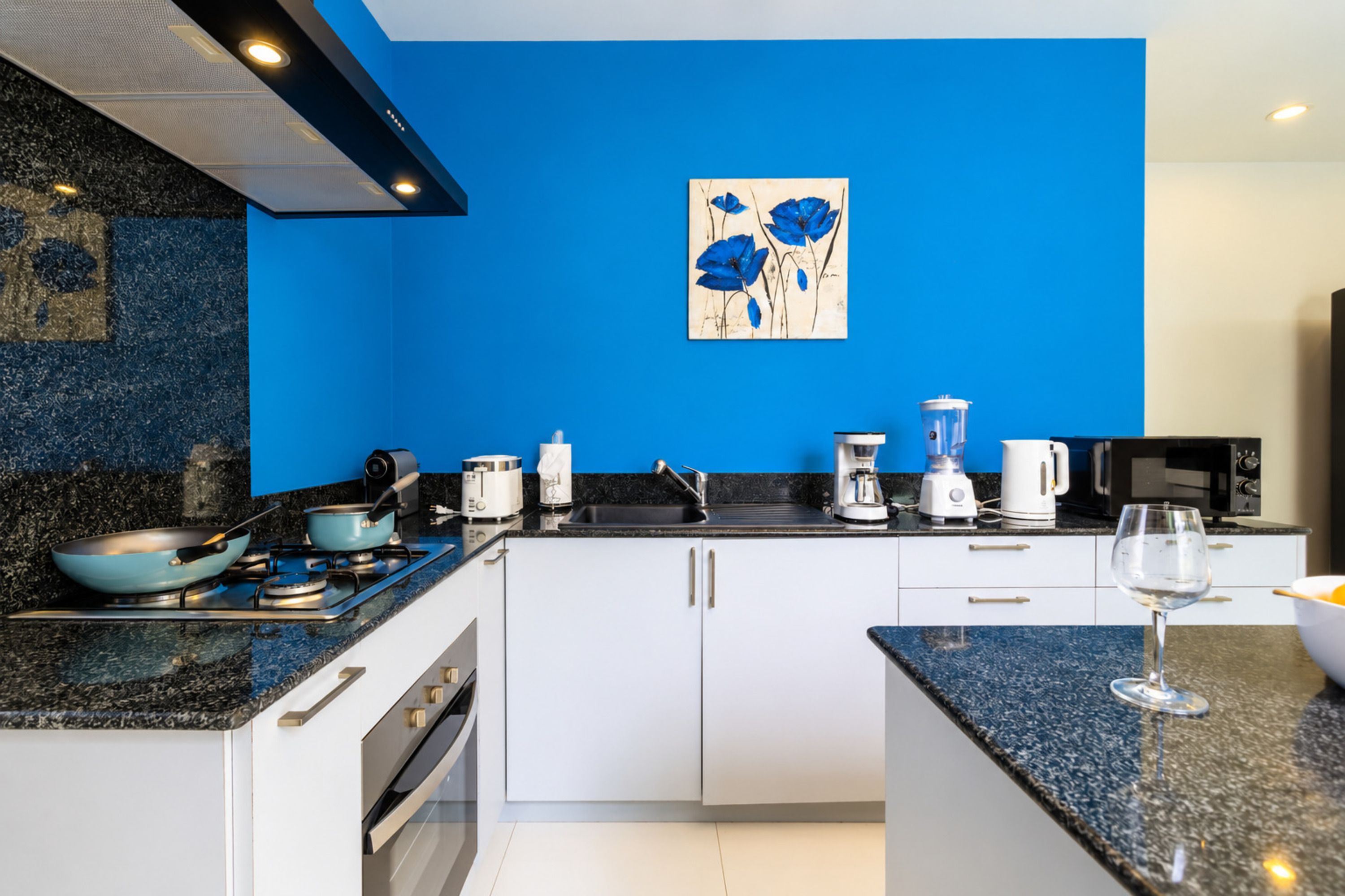 A modern kitchen with white cabinets, a blue backsplash, and a painting of blue flowers on the wall.