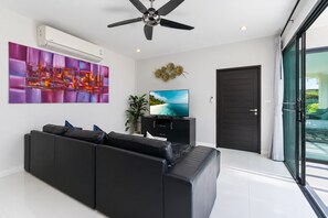 A modern living room with a black sofa, a flat-screen TV, a ceiling fan, and a large window. - 2 BR Pool Villa Clos Vougeot  (Muang Phuket)