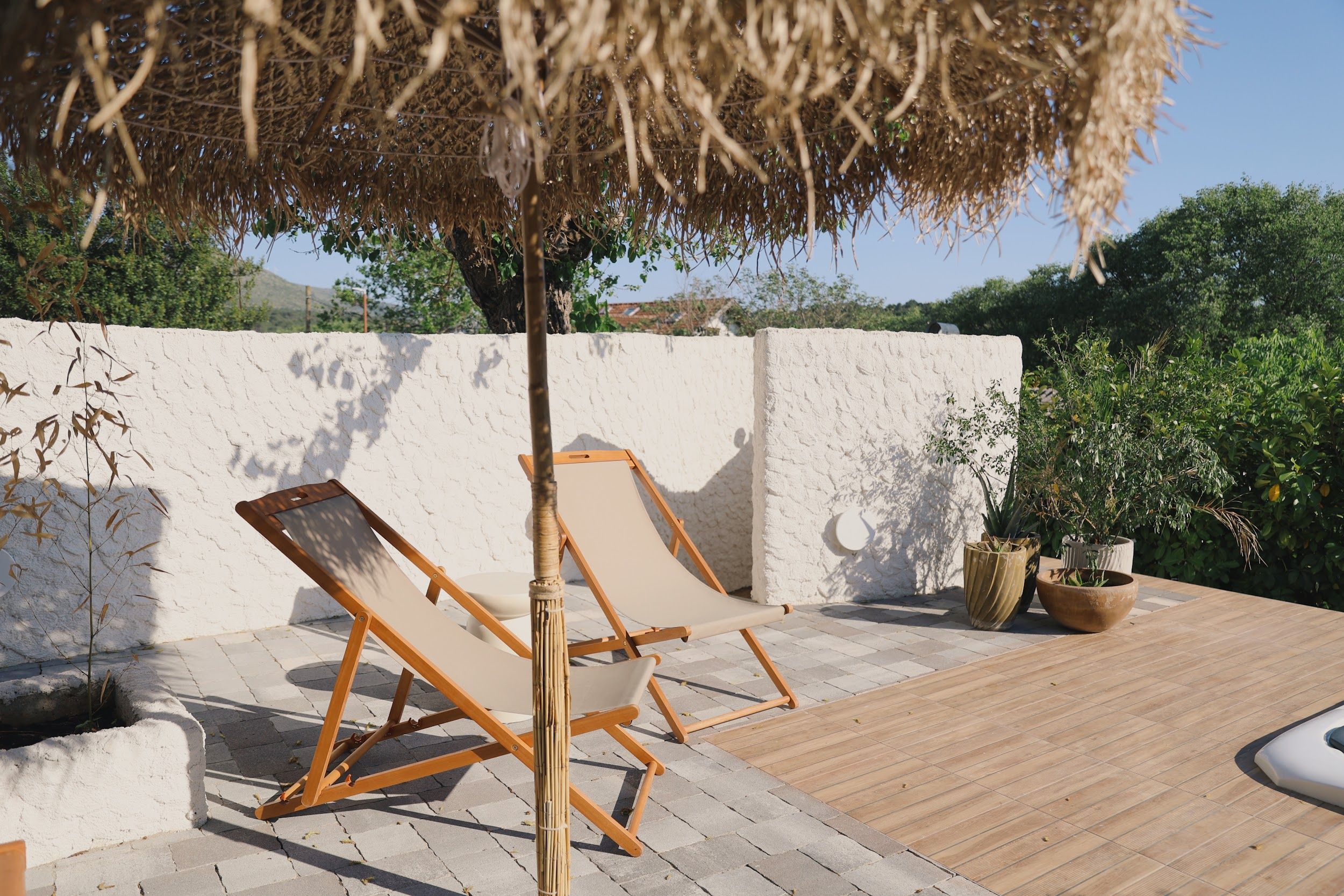 Un espace patio aménagé avec deux fauteuils de jardin en bois, un parasol en chaume et des plantes en pot.