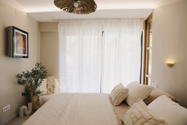 A bedroom with a large bed, a bedside table, a wall-mounted picture, a window with sheer curtains, and a ceiling light fixture.