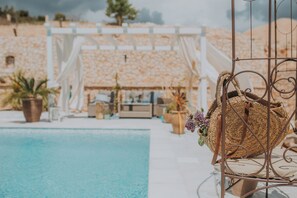 A poolside area with a wicker chair and a metal chair, a white pergola, and a building with a stone facade in the background.