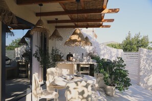 Une terrasse extérieure avec une table à manger, des chaises, des lampes suspendues en chaume et un mur en pierre.