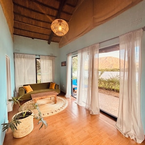 A room with a wooden ceiling, a sofa, a coffee table, a potted plant, and a view through large windows. - Odegaard Lodge (Luangwa Feira)