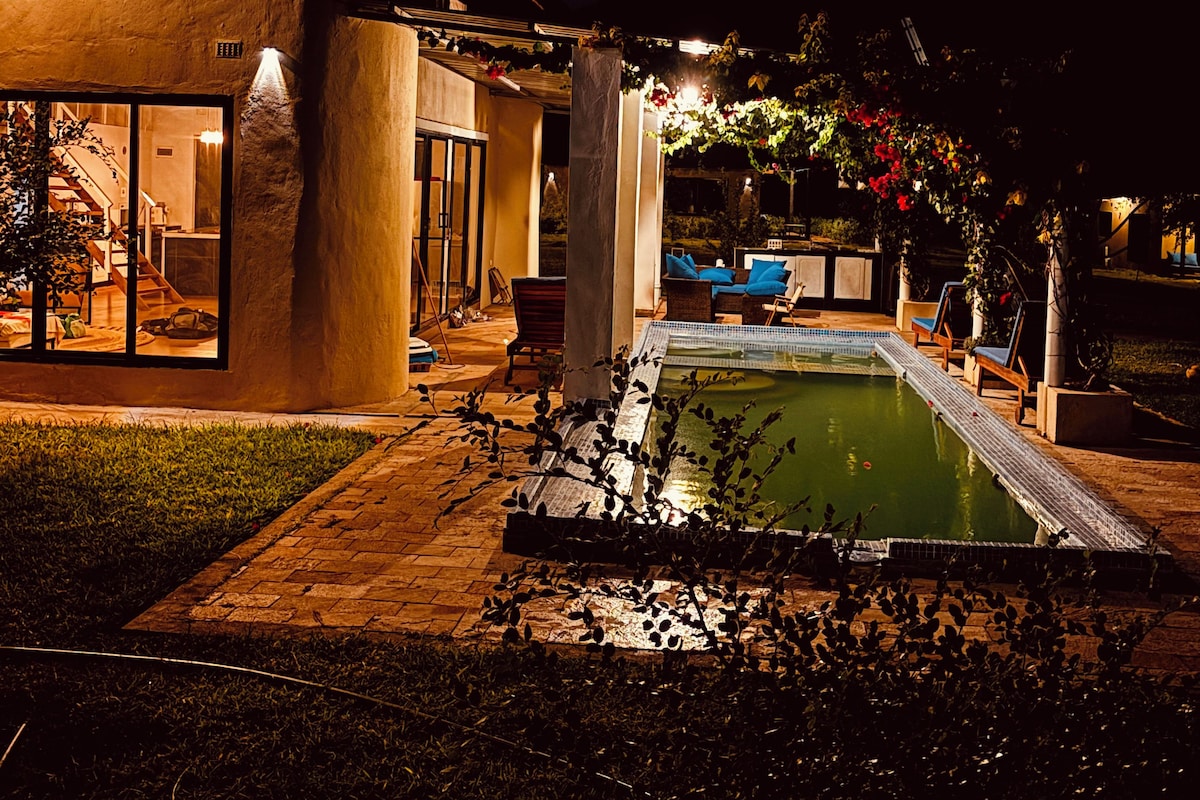 A nighttime view of a modern house with a swimming pool, outdoor seating, and a well-lit patio area.