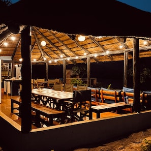 An outdoor dining area with wooden tables and chairs, a thatched roof, and string lights. - Odegaard Lodge (Luangwa Feira)