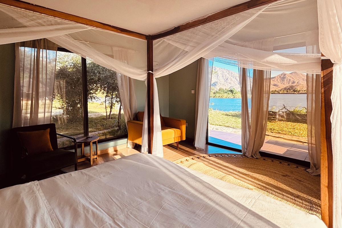 A bedroom with a large bed, a sofa, and a view of a lake and mountains through sheer curtains.
