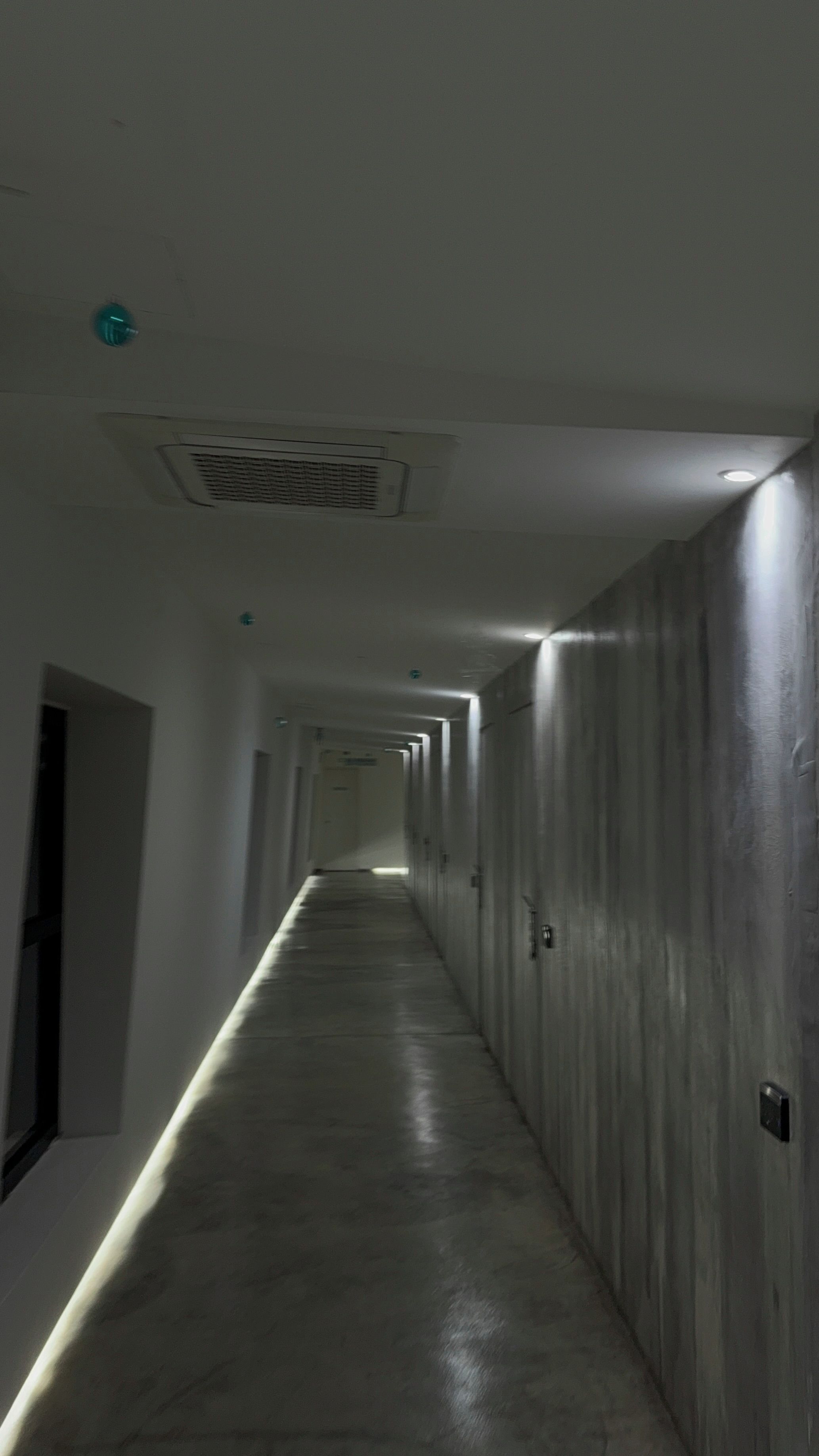 Un lungo e stretto corridoio con diverse porte su un lato e una griglia di ventilazione sul soffitto.