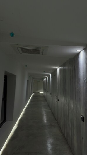 Un lungo e stretto corridoio con diverse porte su un lato e una griglia di ventilazione sul soffitto.