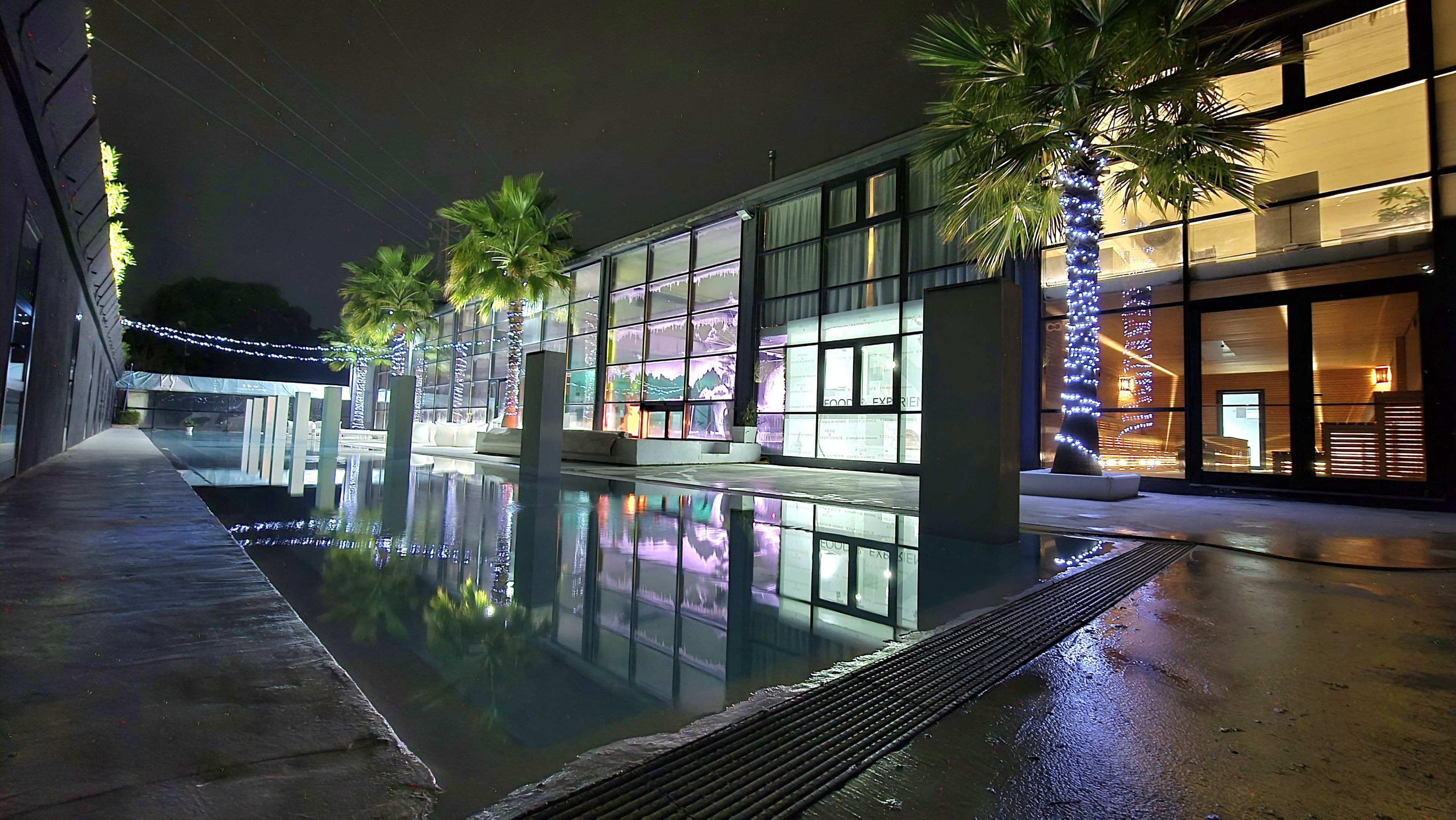 Un espace piscine nocturne avec une barrière en verre, des palmiers et des fenêtres illuminées d’un bâtiment.