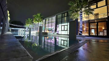 Un espace piscine nocturne avec une barrière en verre, des palmiers et des fenêtres illuminées d’un bâtiment.