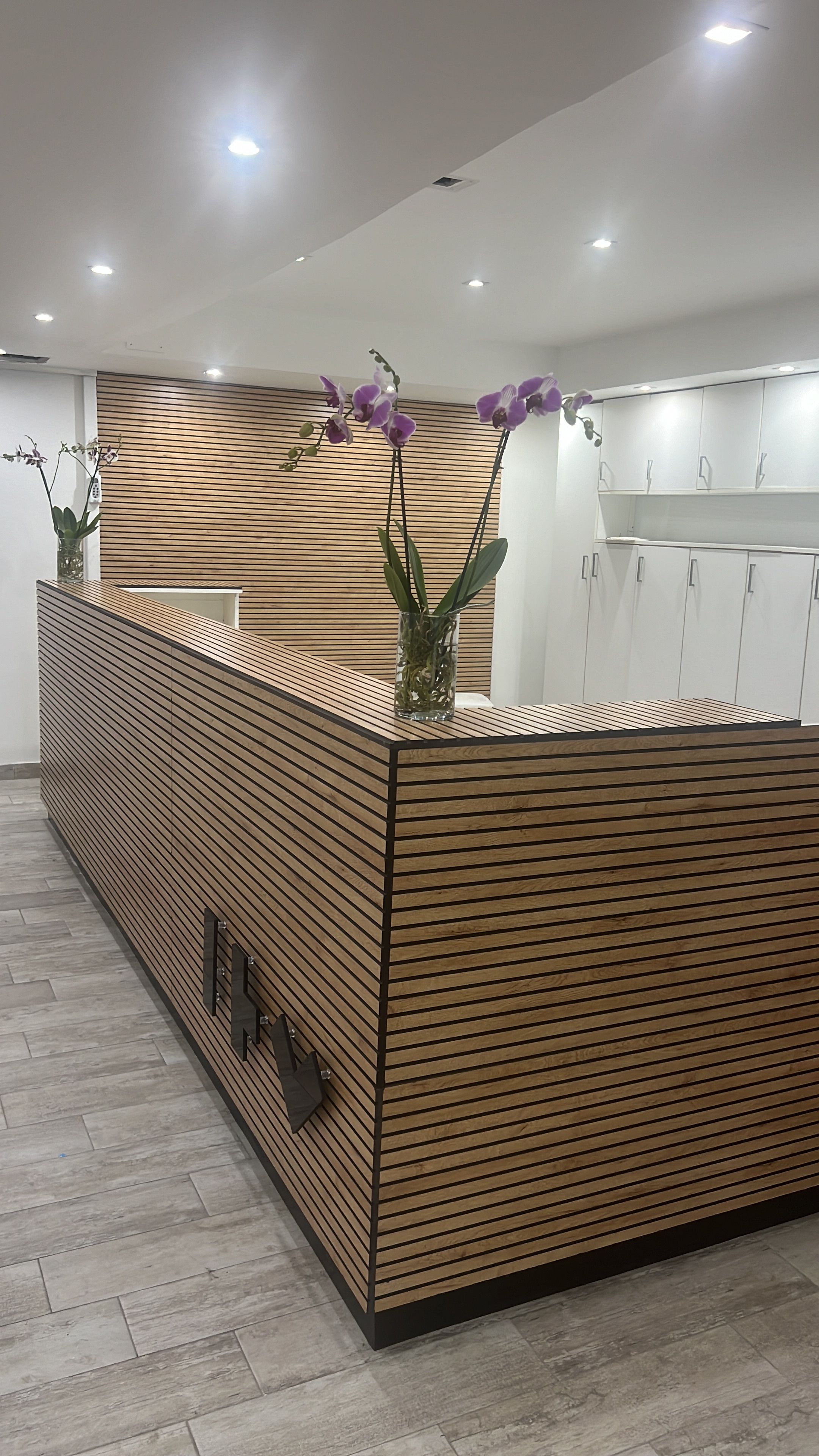 Un comptoir d'accueil avec un design composé de lattes de bois, un vase contenant des orchidées violettes et, au fond, un espace cuisine moderne.