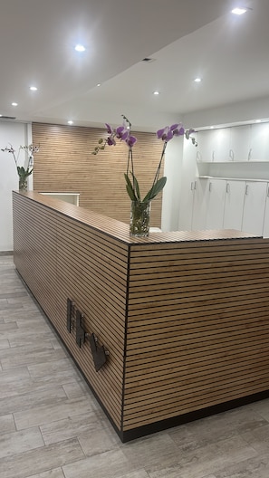 A reception desk with a wooden slat design, a vase with purple orchids, and a modern kitchen area in the background.
