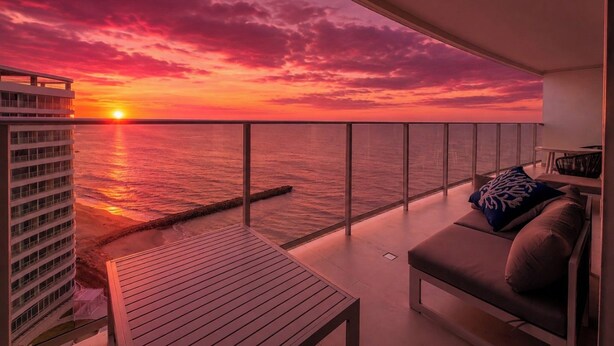 Un balcon offrant une vue sur le coucher de soleil sur l’océan, un coin salon avec un canapé et des coussins, et un immeuble de grande hauteur en arrière-plan.