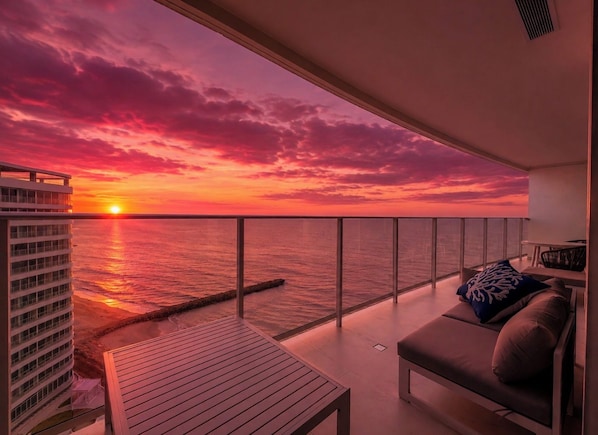 En balkon med udsigt til solnedgangen over havet, et opholdsområde med sofa og puder, og en høj bygning i baggrunden.