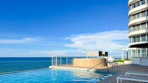 Un espace aménagé au bord de la piscine, avec vue sur l’océan, une terrasse en bois et un bâtiment moderne doté de balcons.