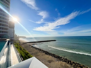 A coastal view with a clear sky, a rocky shoreline, and a building with a balcony overlooking the sea.