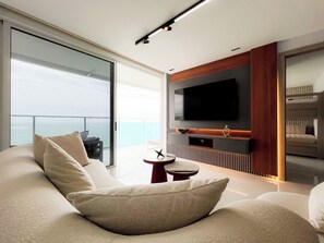 A modern living room with a large flat-screen TV, a sofa, a small table, and a view of the ocean through large glass doors.