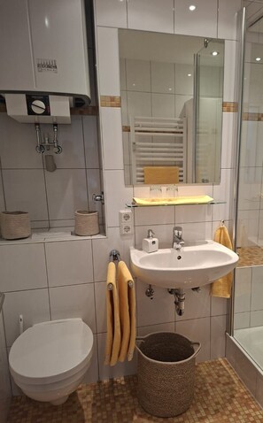 Ein modernes Badezimmer mit Toilette, Waschbecken und Dusche. Handtücher und ein Mülleimer sind vorhanden.