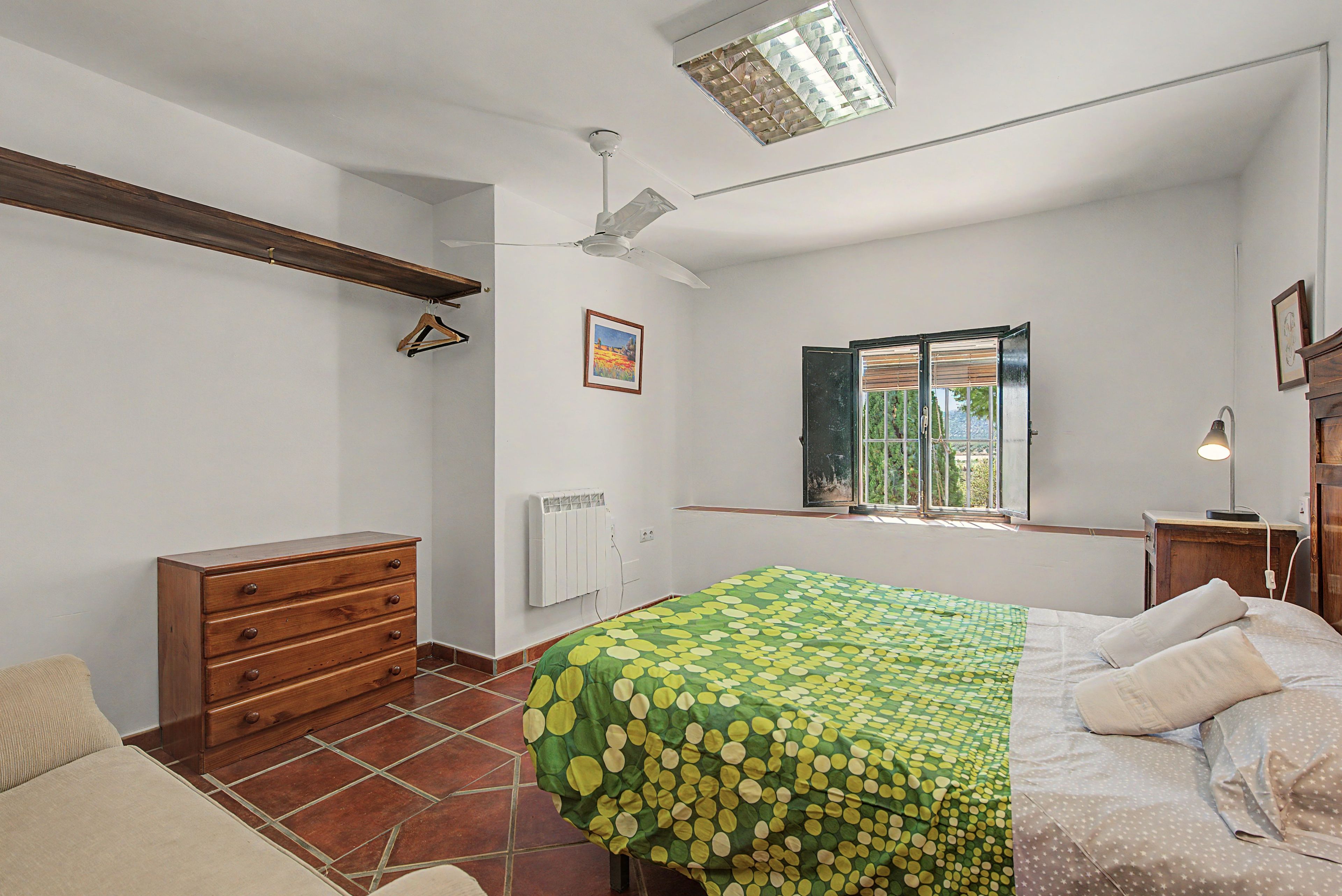 Un dormitorio con cama, cómoda, radiador, ventana con persianas y ventilador de techo.