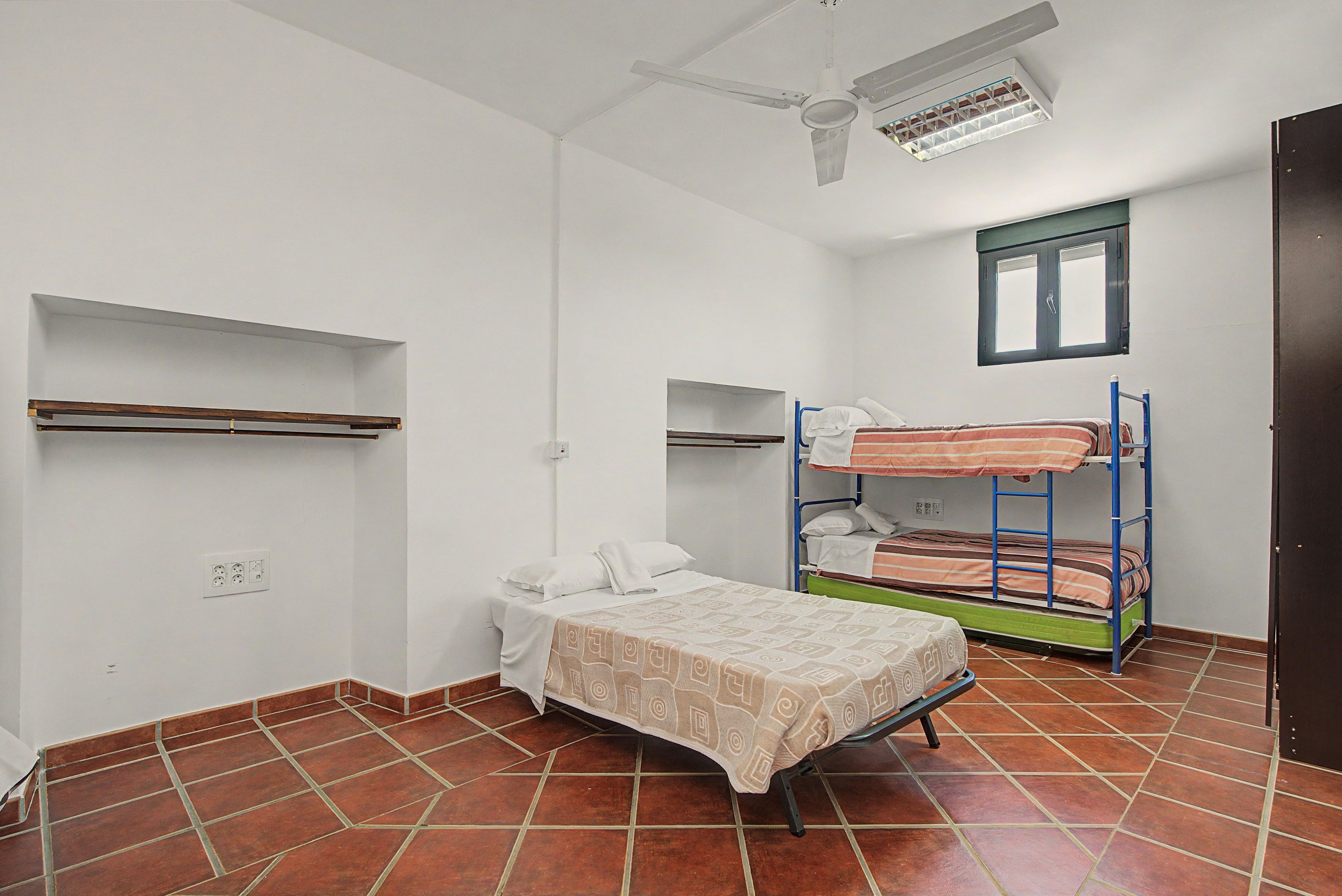 A room with a bunk bed, a single bed, a ceiling fan, a window, and a door.