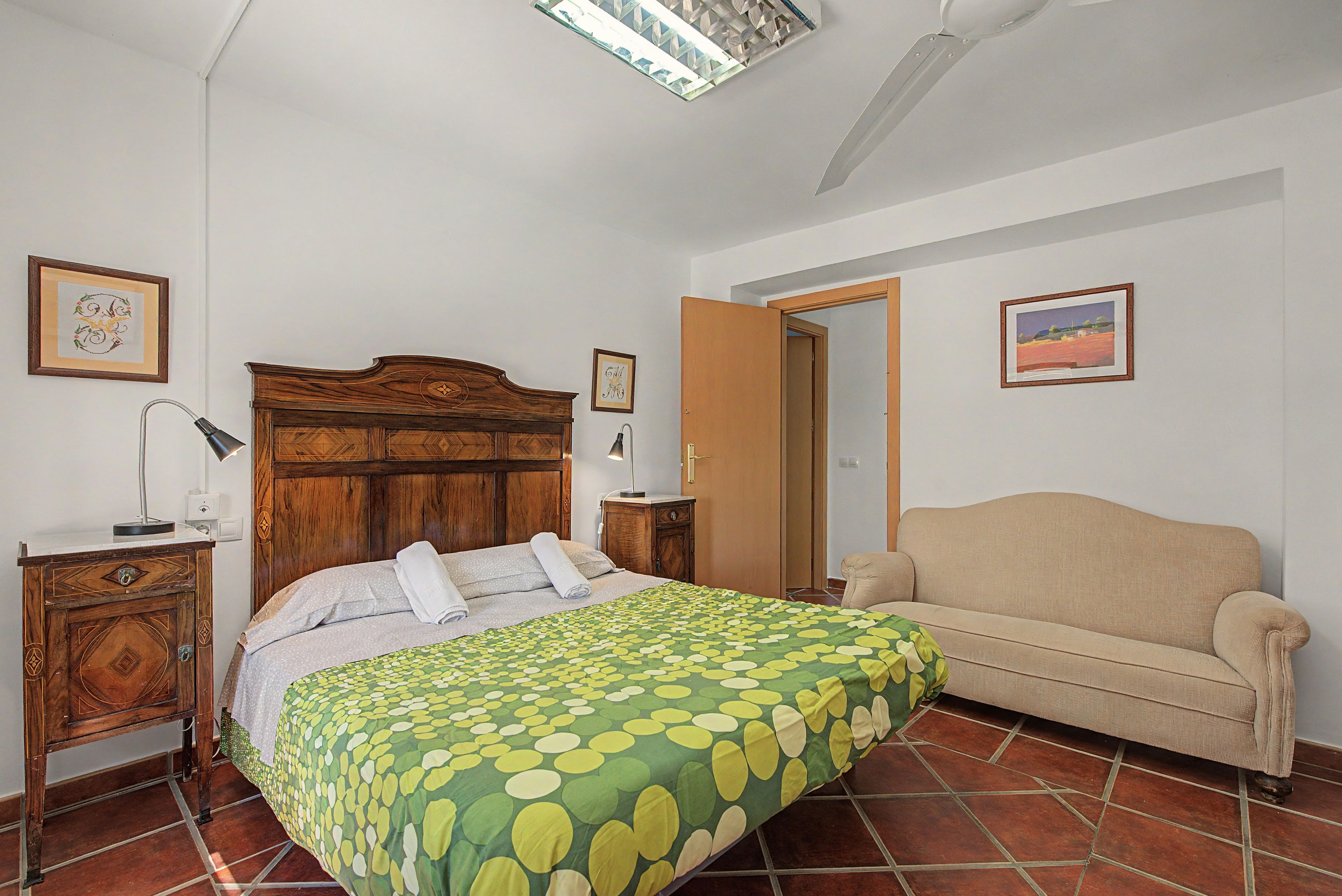 Un dormitorio con cabecera de madera, colcha de cama verde con lunares, sofá color beige y piso de baldosas.