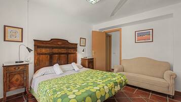 Un dormitorio con cabecera de madera, colcha de cama verde con lunares, sofá color beige y piso de baldosas.