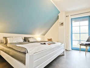 A bedroom with a bed, pillows, a bedside table, a chair, and a window with a blue door. - 11a/t6 Holiday Home Park \"freesenbruch\" (Zingst)