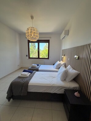 A bedroom with two beds, a window with a view of greenery, a hanging light fixture, and a wall-mounted air conditioning unit. -  Spacious 6-bedroom house in beautiful Poli Chrysochous with AC (Poli Chrysochous)
