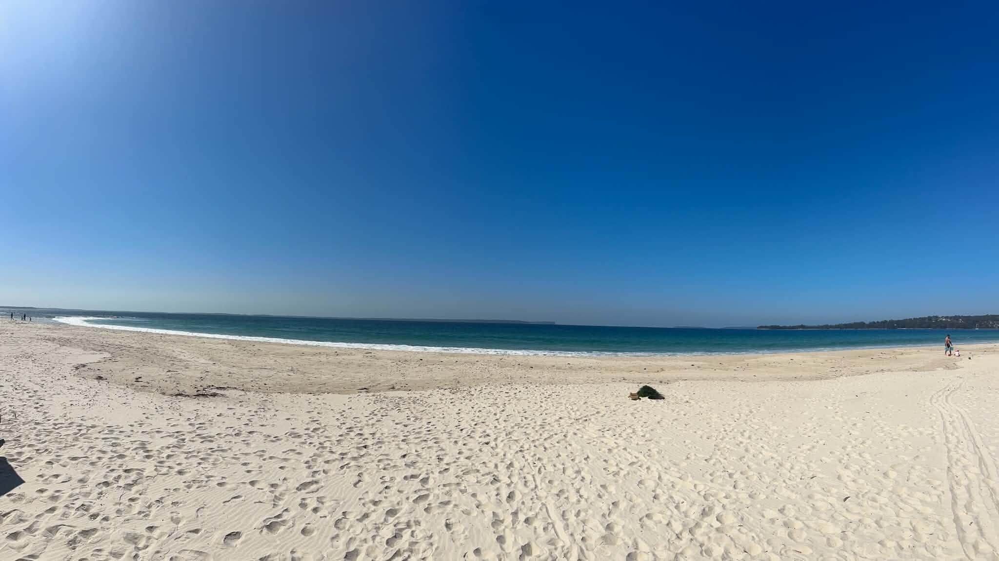 A wide sandy beach with footprints leading to the water, a clear sky, and a distant shoreline.