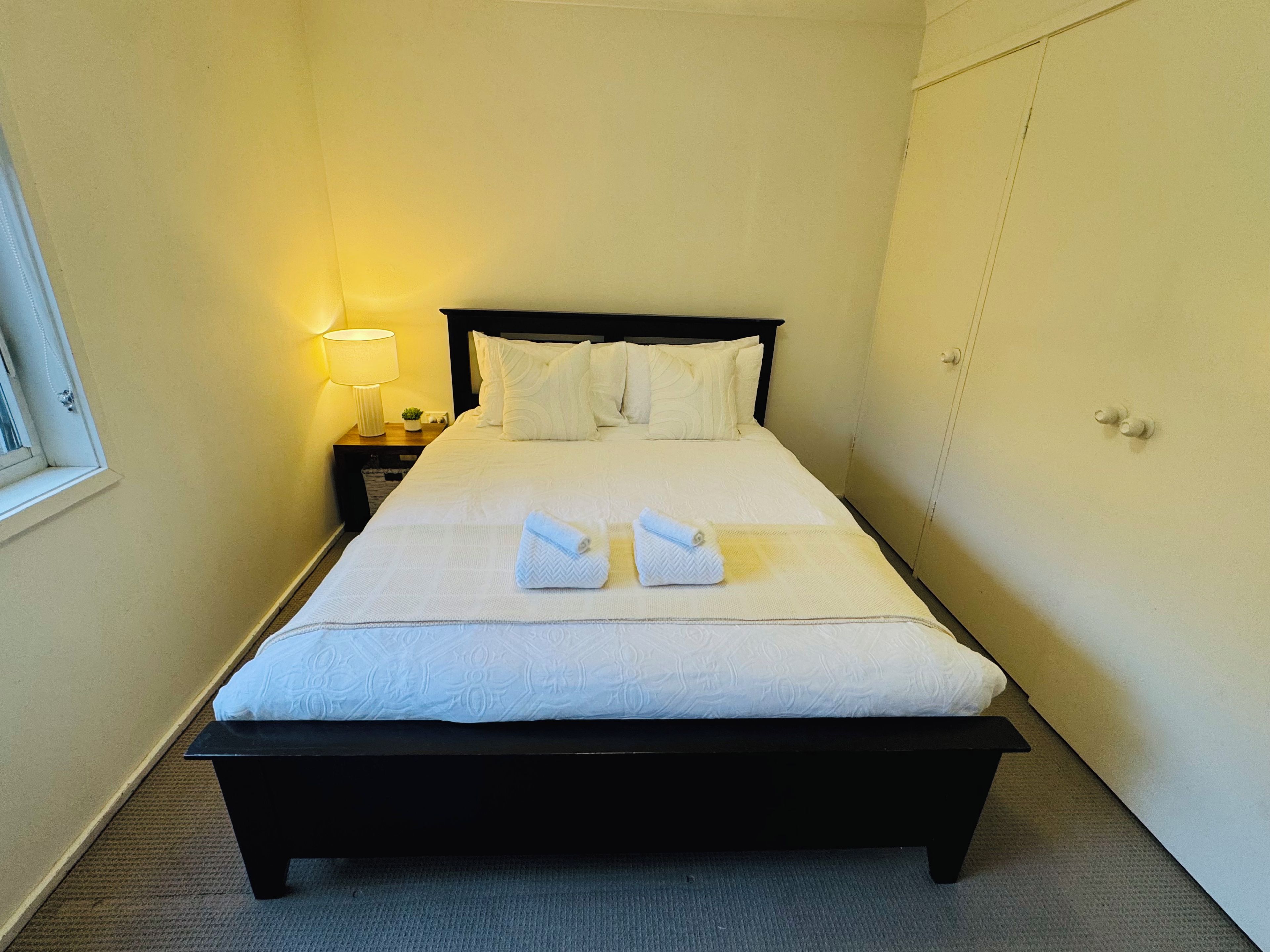 A neatly made bed with white linens and two towels folded on top, a bedside lamp, and a window on the left side of the room.