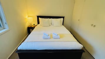 A neatly made bed with white linens and two towels folded on top, a bedside lamp, and a window on the left side of the room.