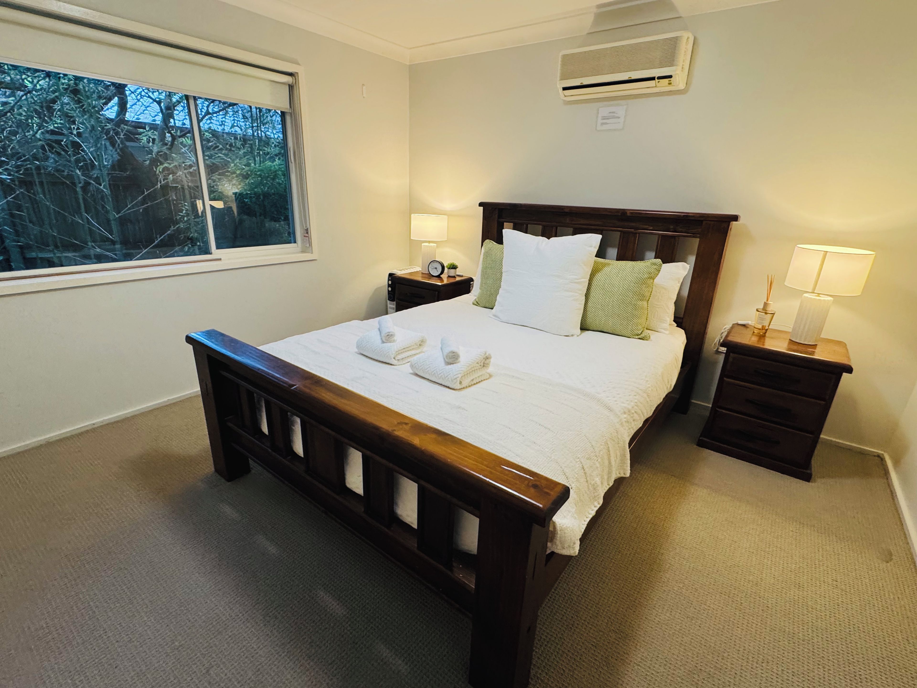 A neatly made bed with white linens and two towels, a bedside table with a lamp, and a window with a view of trees.