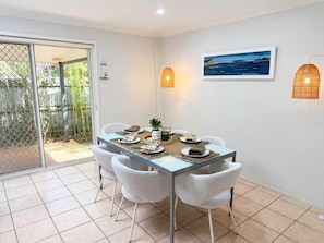 A dining area with a table set for four, chairs, a picture on the wall, and a view through a sliding door. - Samson Sea-clusion 3 bedroom home in Vincentia (Vincentia)