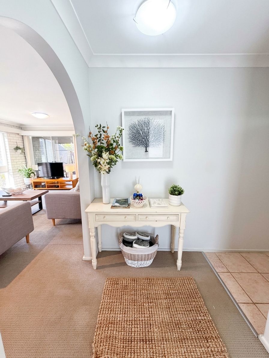 A living room with a white console table, a framed artwork, a vase with flowers, and a television in the background.