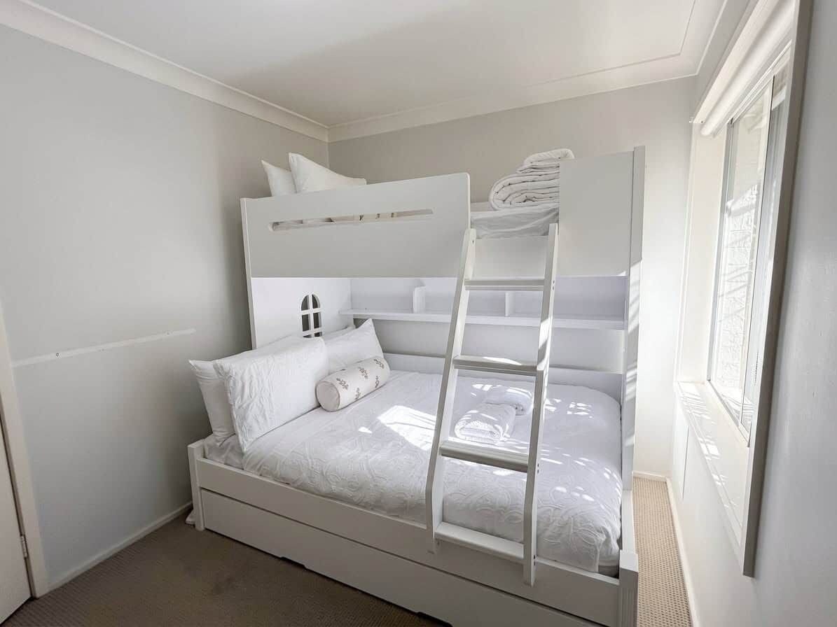 A bunk bed with a ladder, white bedding, and a window with blinds.