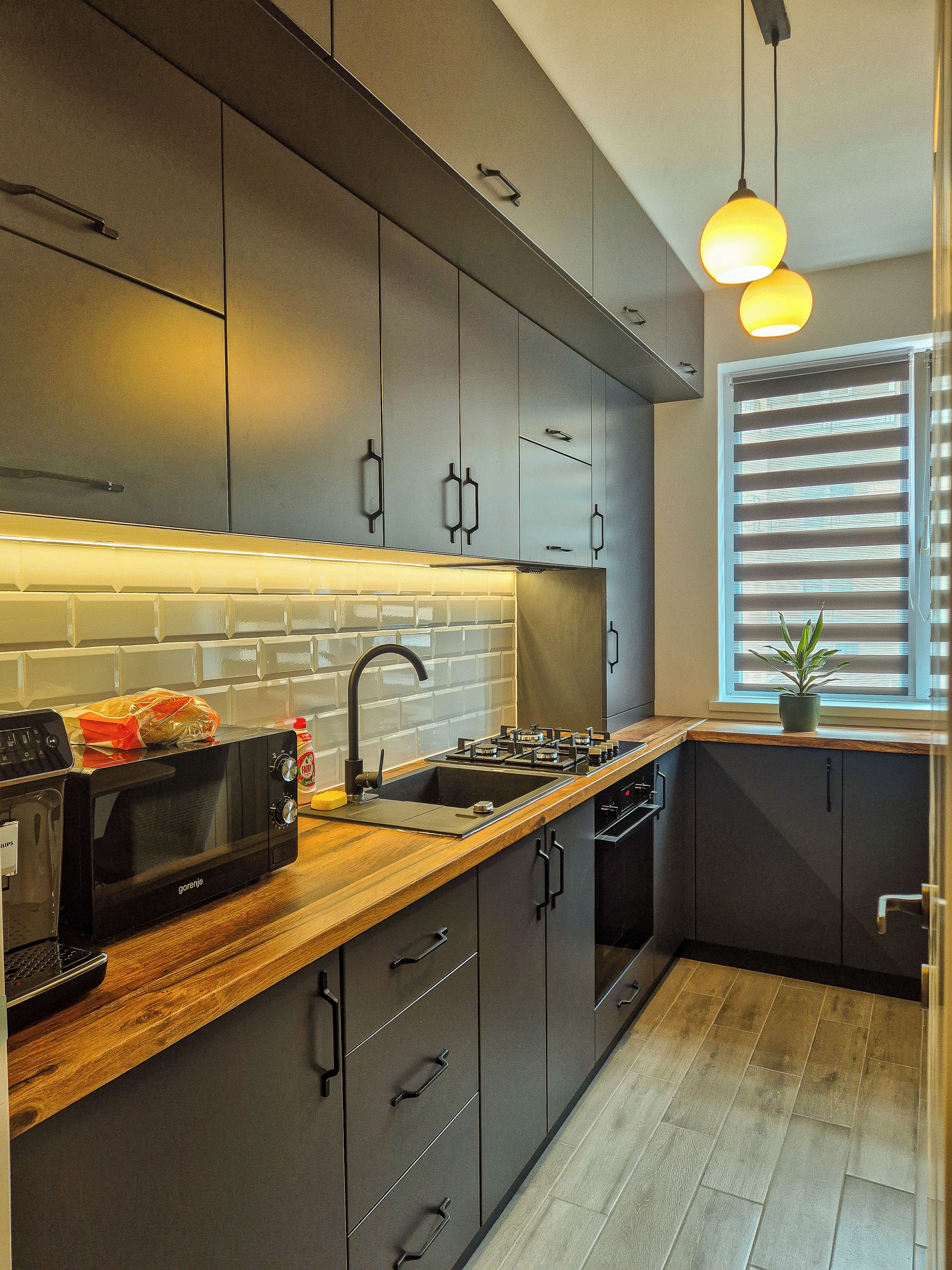 A modern kitchen with dark cabinetry, a wooden countertop, a built-in microwave, and a window with blinds.