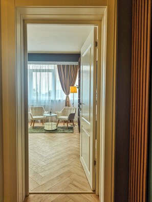 A hotel room with a herringbone floor, a seating area with two armchairs and a small table, and a view through a door to a balcony with curtains.