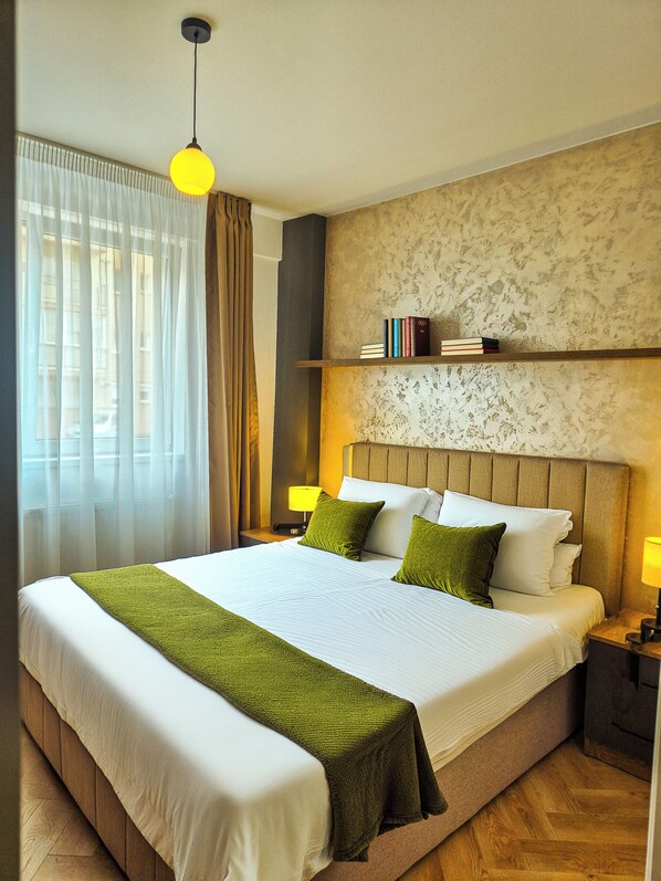 A hotel room with a large bed, a bedside lamp, a nightstand, a bookshelf, and a window with curtains.