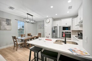 A modern kitchen with white cabinetry, a stainless steel refrigerator, a microwave, and a gas stove. There is a dining area with a wooden table and chairs, and a large window with blinds. - Reunion Village 4BR Mickey and Moana Retreat (Kissimmee)