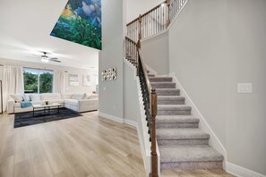 A spacious living room with a large window, a staircase with a wooden railing, and a ceiling mural. - Reunion Village 4BR Mickey and Moana Retreat (Kissimmee)