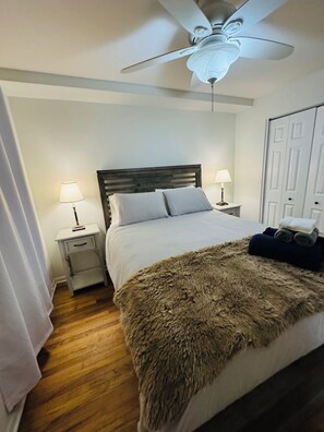 Een netjes opgemaakt bed met een zachte deken, twee bedlampen, een plafondventilator en een houten vloer.