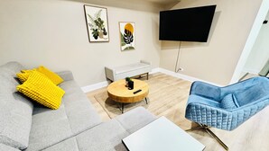 En moderne stue med en grå sofa, gule puder, et rundt træ sofabord, en blå lænestol og et fladskærms-tv monteret på væggen.