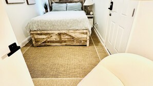 A bedroom with a wooden bed frame, a white chair, a bedside table with a lamp, and a door.
