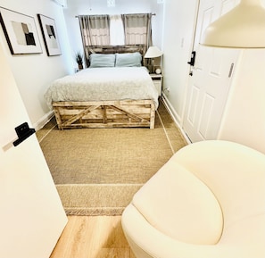 A bedroom with a wooden bed frame, a white chair, a bedside table with a lamp, and a door.