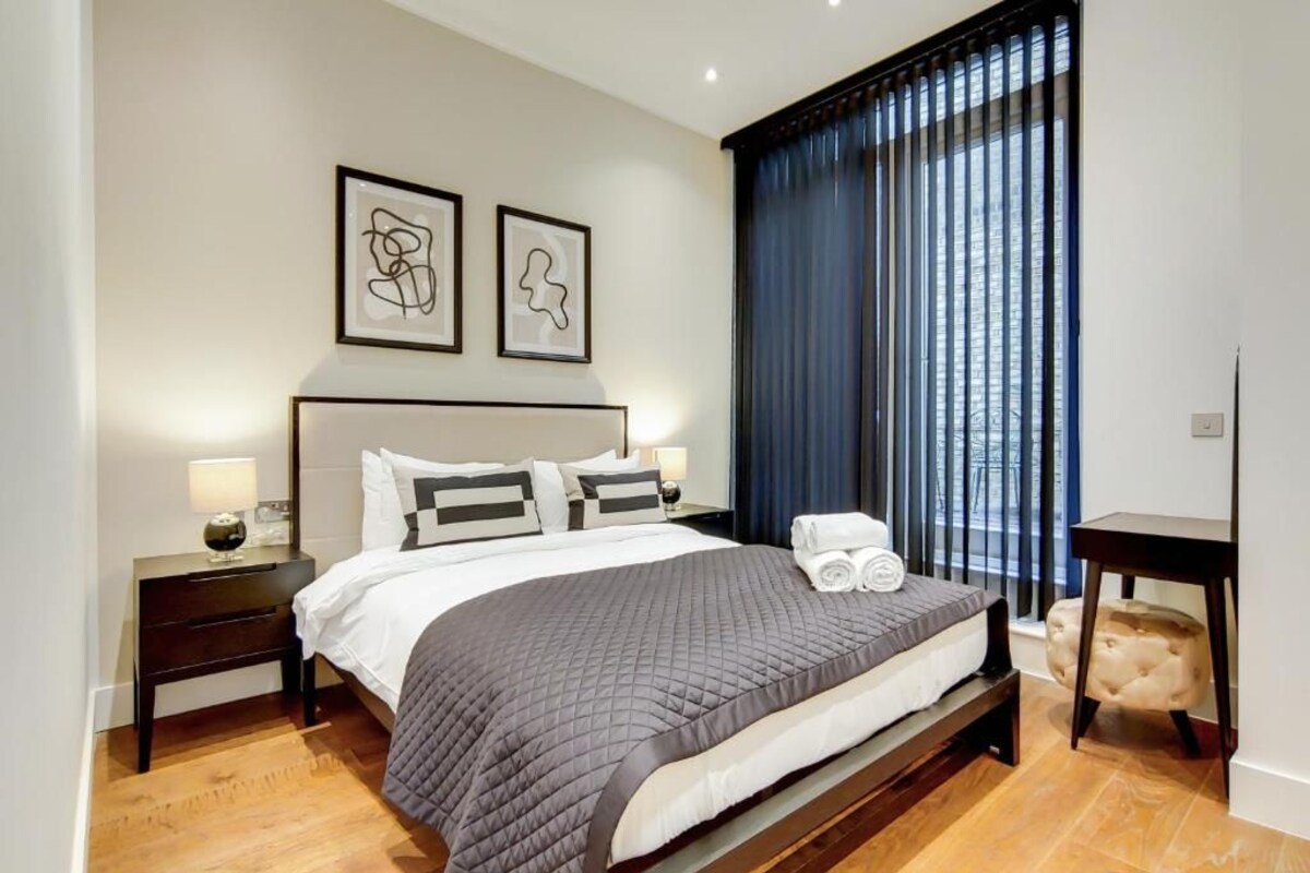A neatly made bed with a grey quilt, two bedside tables with lamps, and a window with vertical blinds.